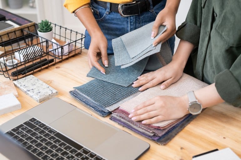 Designer selecting fabric swatches on wooden table with laptop showing fabric comparison for clothing projects