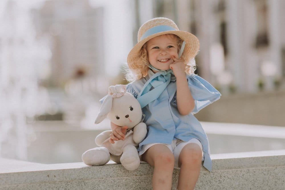 Smiling toddler in a straw hat and a blue dress plays with a stuffed rabbit outdoors