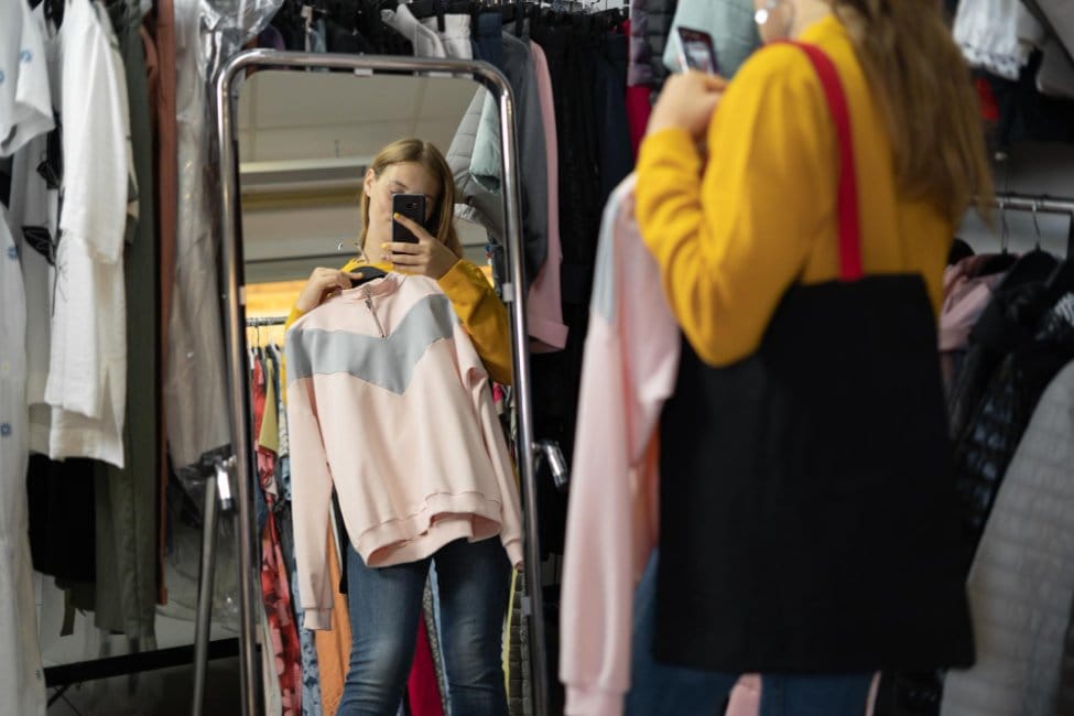A woman standing in front of a mirror in a clothing store holding an outerwear and taking pictures with a phone