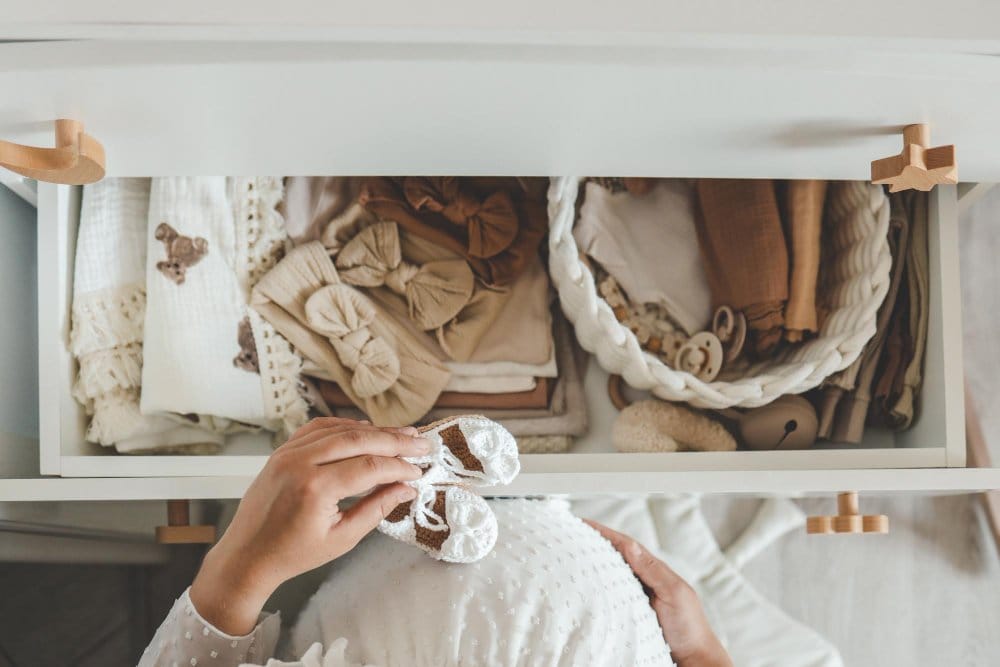Pregnant woman standing in front of a drawer full of baby and toddler clothing and holding a handmade baby shoe