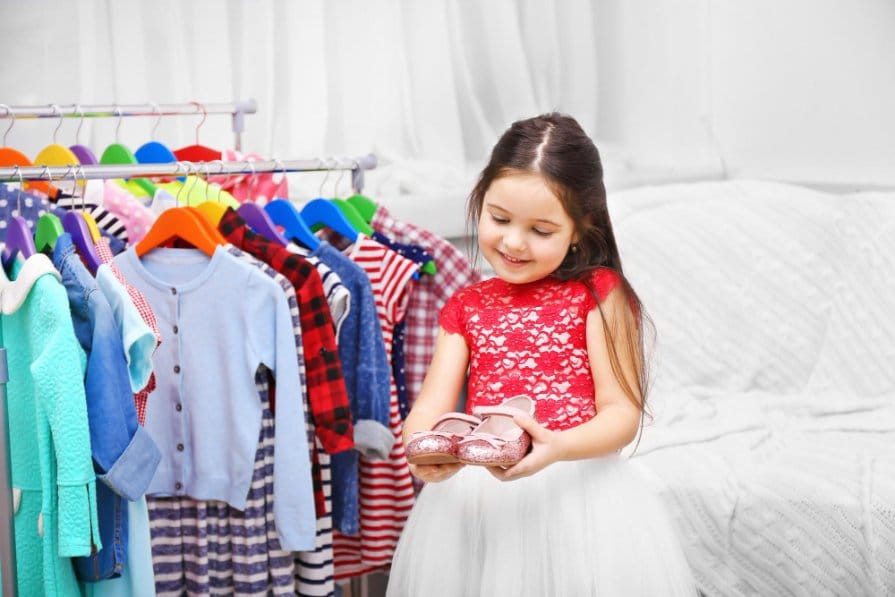 Little girl standing in front of a clothing rack and holding shoes in her hands