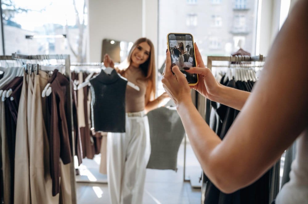 two female friends in a shop, one of them holding a clothing item and the other one holding a mobile phone doing live video with the other friend