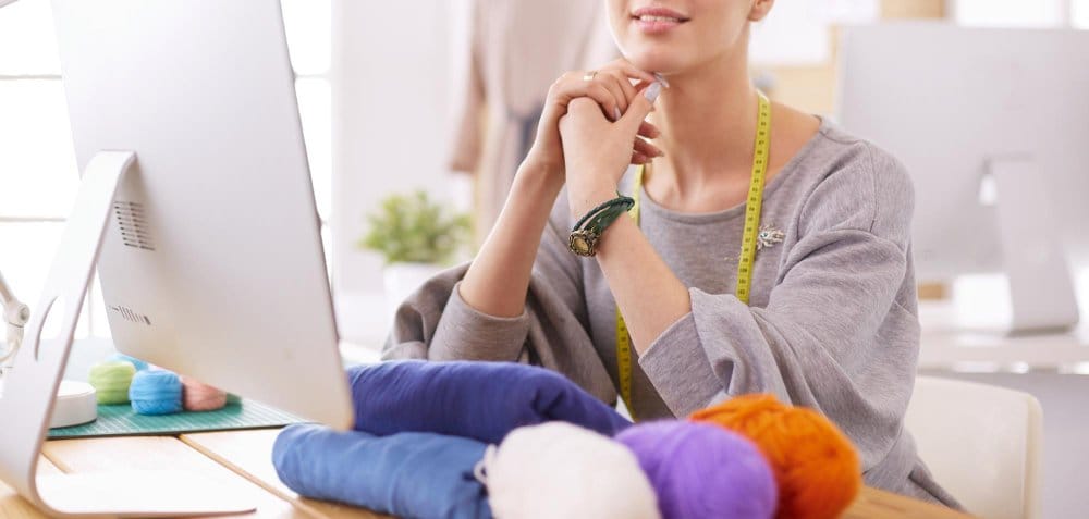 A fashion designer sitting in front of a computer with folded fabrics and knitting thread on the table