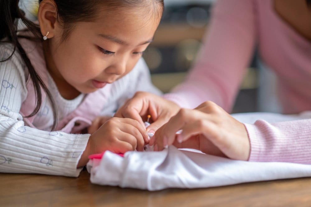Child learning to sew button with adult guidance at table