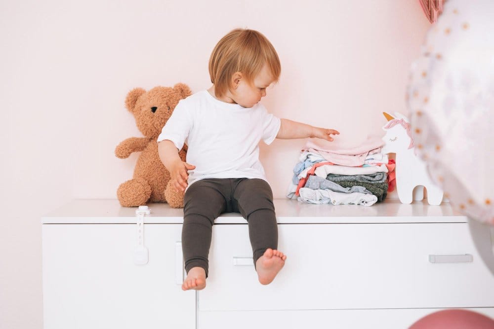 little girl sitting on a white drawer with folded clothes and teddy bear