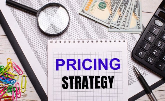 On the office table there is a notebook with the text PRICING STRATEGY, a pen, a calculator, dollar notes, multi-colored paper clips and a magnifying glass