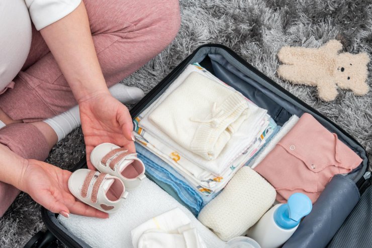 Pregnant woman sitting on the floor holding toddler shoes on top of packed suitcase readying for travel