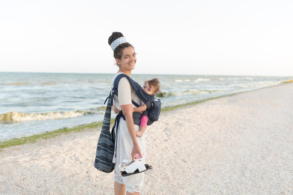 A mother standing on a beach carrying her toddler with a baby carrier
