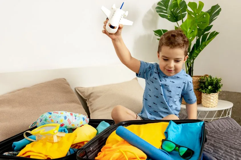 Travel accessories for toddlers including shoes, hats, bibs, and comfort items arranged alongside a packed suitcase