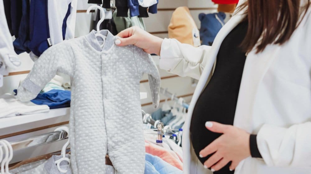 Pregnant woman holding a gray baby onesie in a store while considering infant sizing factors