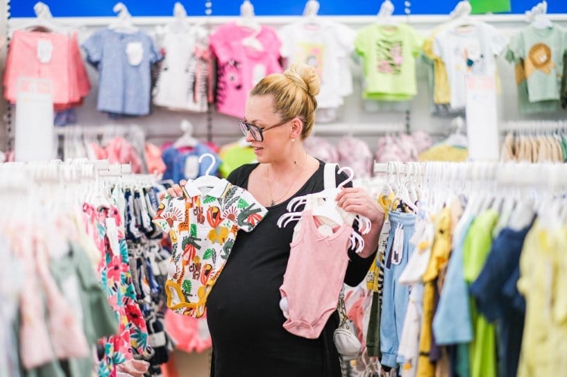 Expectant mother comparing two baby bodysuits of different sizes in a retail store