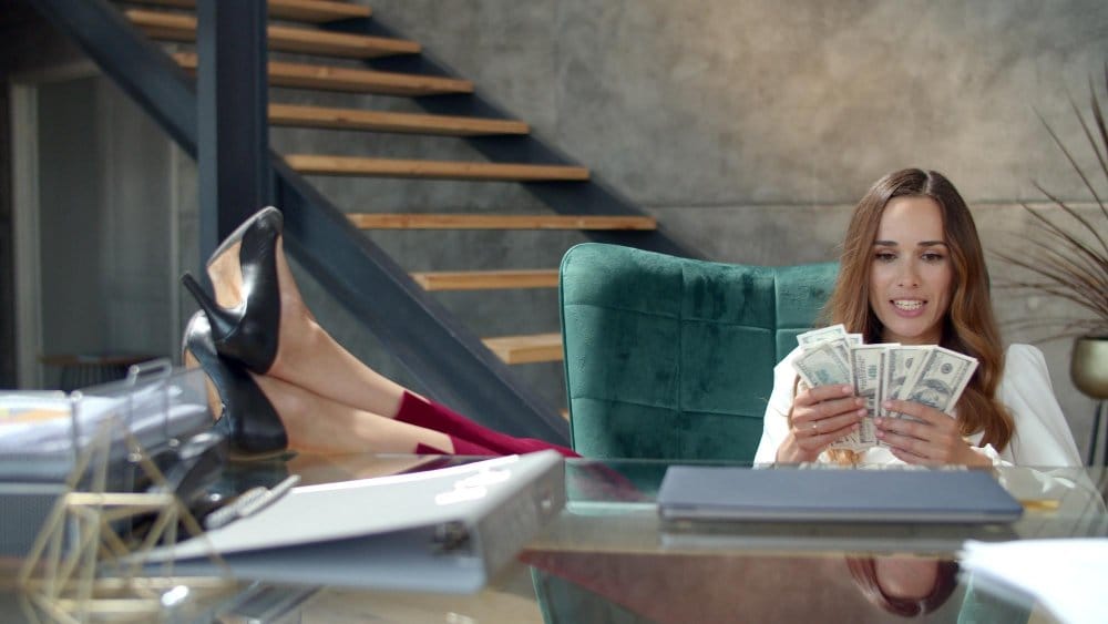 A woman sits in a green velvet chair behind a glass desk, counting a stack of US dollar bills with a pleased expression, probably after making sales on either vinted or ebay