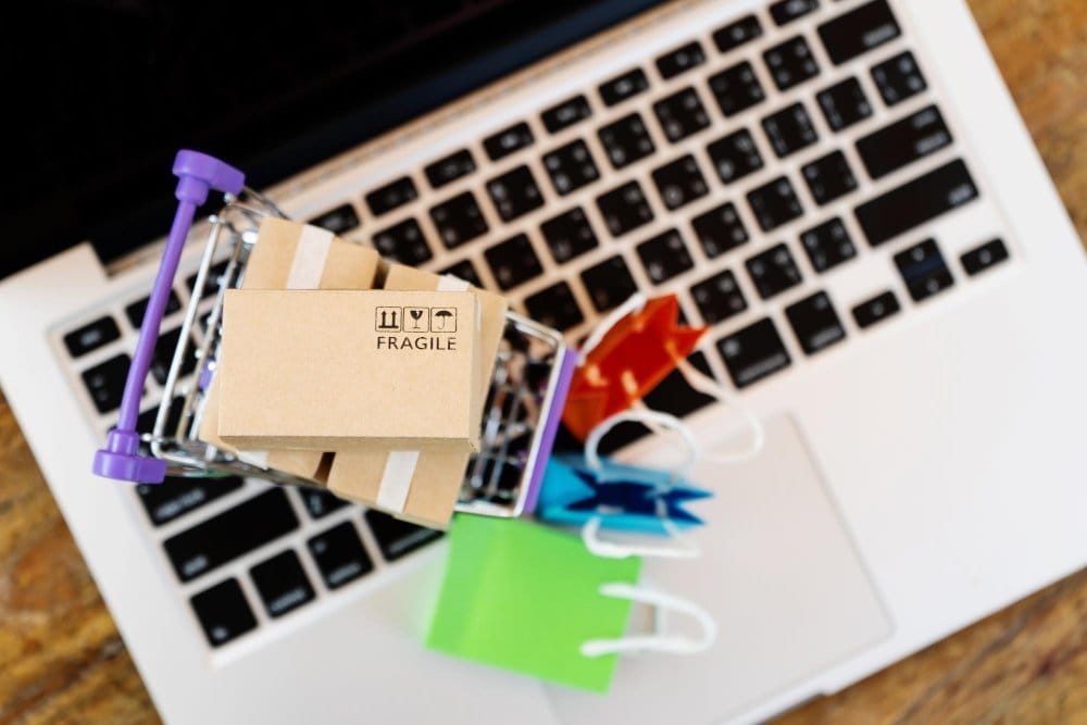 A miniature shopping cart containing small cardboard boxes labeled "fragile" is placed on a laptop keyboard, with colorful shopping bags nearby, representing ebay and vinted