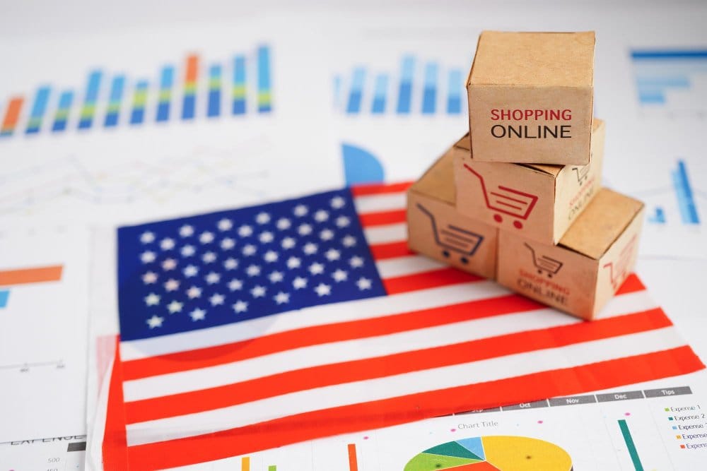 Small cardboard boxes labeled "Shopping Online" stacked on top of an American flag, surrounded by various colorful business charts and graphs in the background.