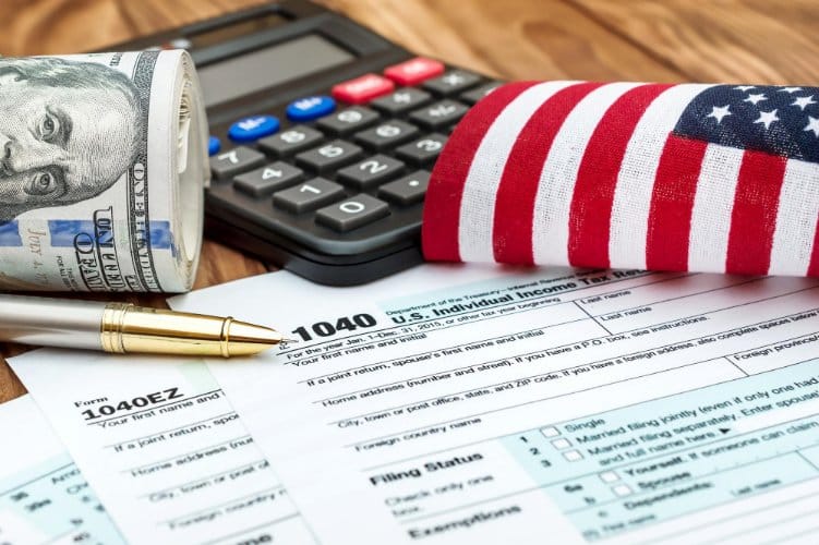 Close-up of a U.S. Individual Income Tax Form 104 with a pen, a rolled-up hundred-dollar bill, a calculator, and a folded American flag on a wooden surface.