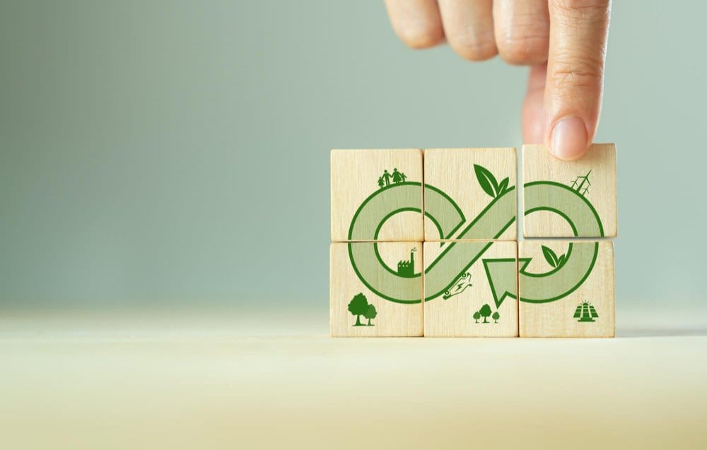 A hand is placing the last wooden block in a set of six blocks that together form a green circular arrow symbolizing a sustainable and circular economy. The blocks feature small icons representing nature, industry, and renewable energy, emphasizing environmental sustainability