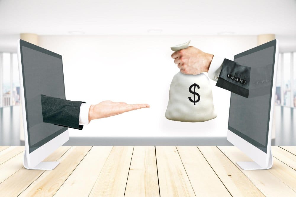 Two computer monitors facing each other on a wooden table, with a hand extending from the left screen offering an open palm, and a hand extending from the right screen holding a money bag with a dollar sign, symbolizing ebay and vinted financial transactions or digital money exchange.