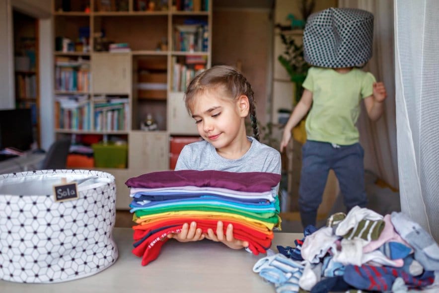 Little girl folding old clothes into pile and her sibling playing in the background