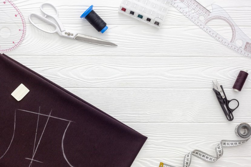 Flat lay of sewing tools including scissors, thread, chalk, rulers, fabric with marked patterns, and a measuring tape on a wooden surface.