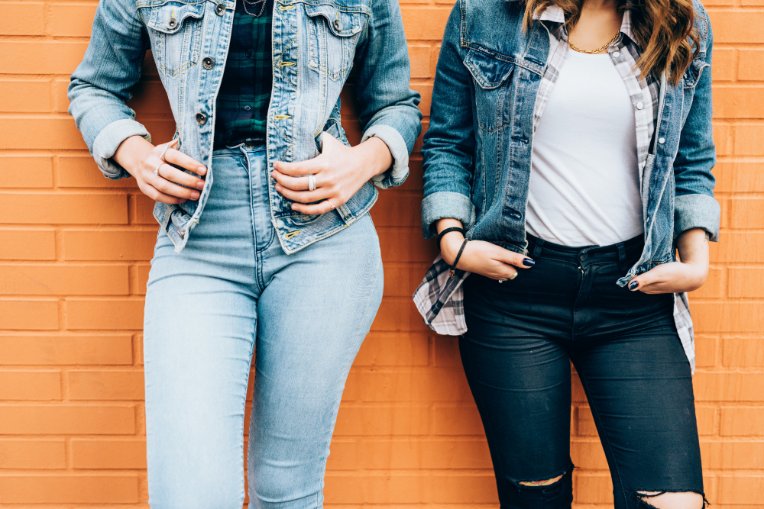 Two women wearing denim jeans and jackets standing by a wall.