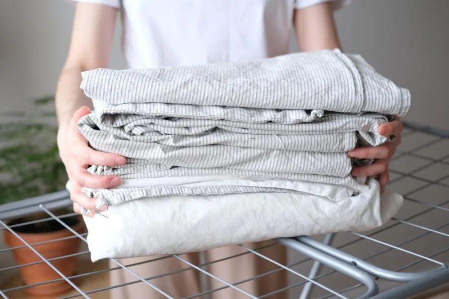 Caring for Stonewashed Linen Sheets Person holding stack of freshly laundered stonewashed linen sheets in gray and white stripes demonstrating proper linen care