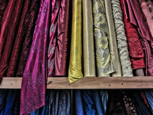 Rows of Banarasi silk fabric rolls in various colors and textures displayed on a wooden rack