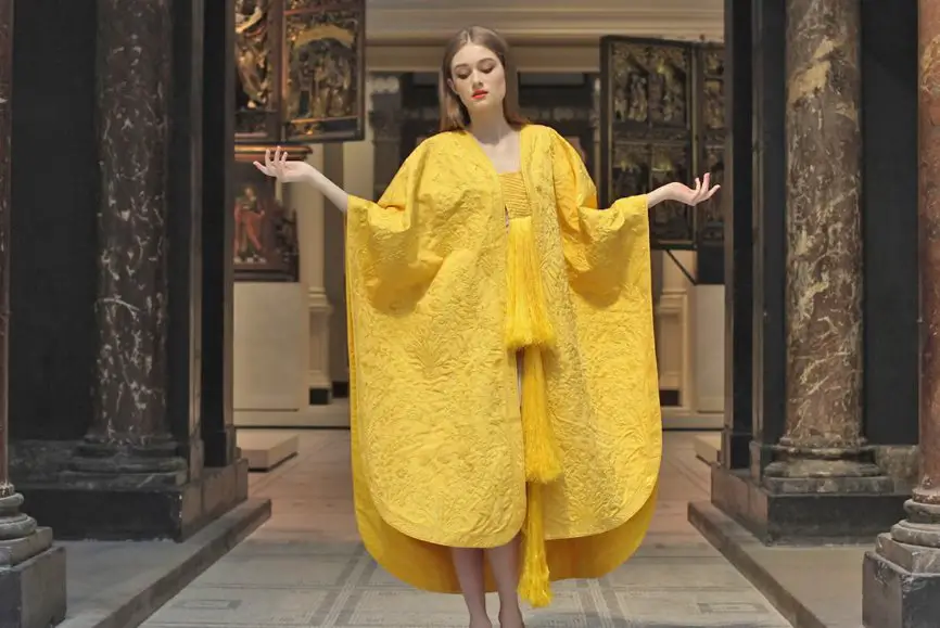 Model wearing a bright yellow spider silk garment with wide sleeves and textured embroidery, standing in a museum like interior