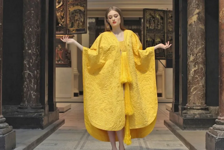 Model wearing a bright yellow spider silk garment with wide sleeves and textured embroidery, standing in a museum like interior