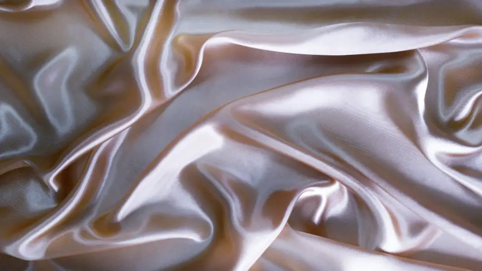 Soft pink charmeuse fabric with a glossy surface arranged in flowing folds that highlight its luxurious sheen