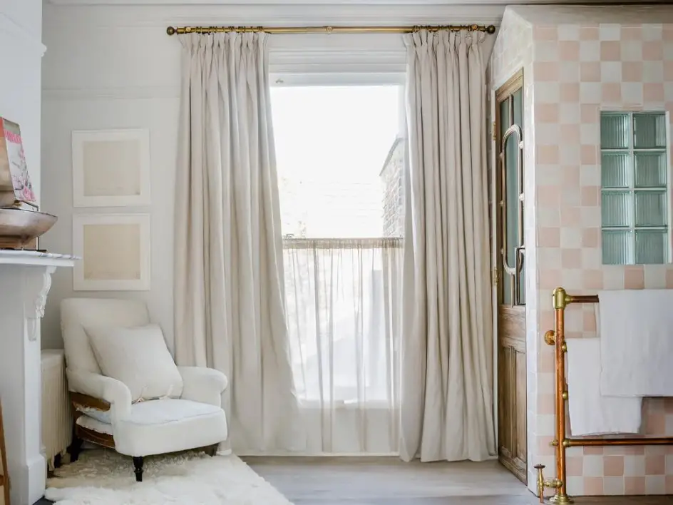 Living room with cream colored tasar silk curtains hanging from a brass curtain rod beside a cozy armchair