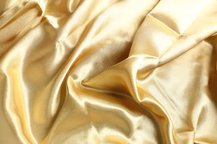 Gold antique satin fabric draped in soft folds showing its textured surface and subtle sheen.