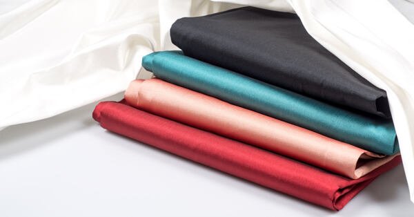 Folded art silk fabrics in red, peach, teal, and black shades displayed with a white cloth backdrop