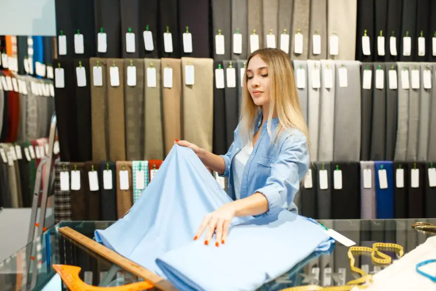 Woman examining light blue fabric in a store surrounded by shelves of textiles