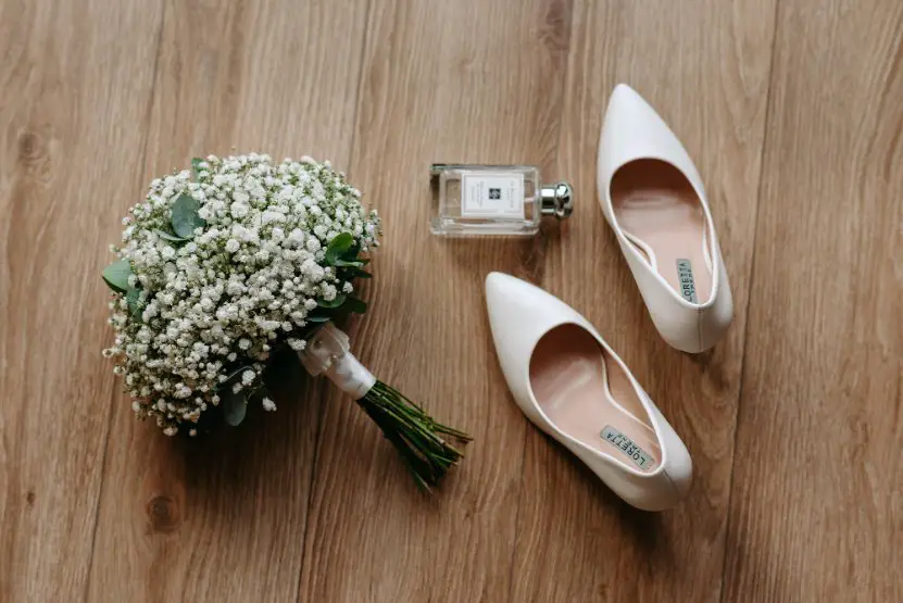 White bridal shoes, bouquet of baby's breath flowers, and perfume arranged on a wooden floor