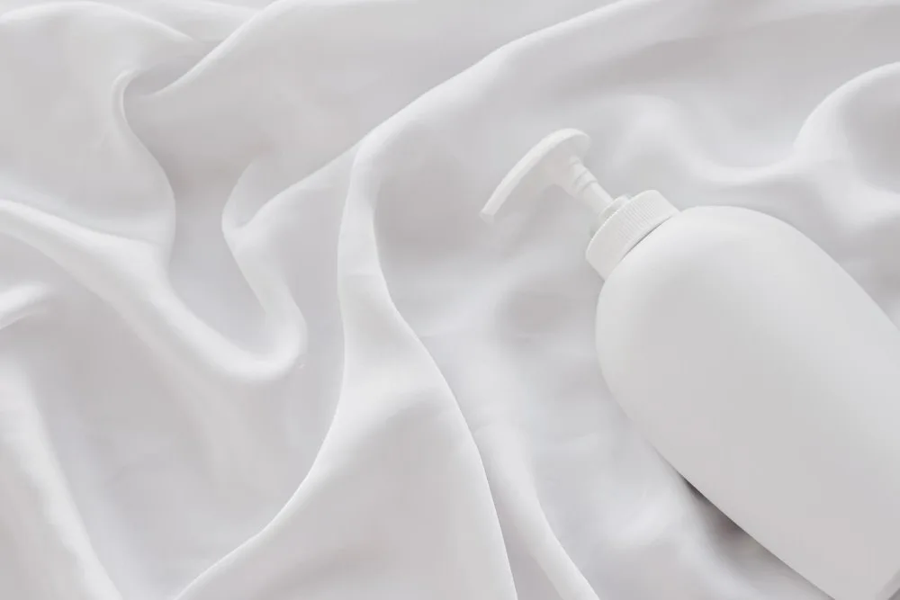 White bottle of fabric care product placed on white acetate satin fabric to represent cleaning and maintenance steps