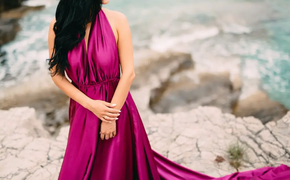 Woman wearing a vibrant magenta silk satin dress standing near the ocean with flowing fabric and soft drape