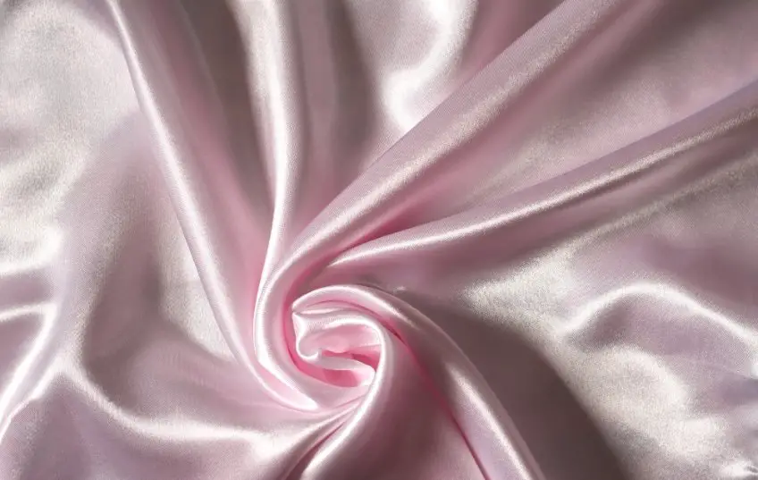 Close up of pale pink satin fabric twisted into soft folds that emphasize its shine and woven structure