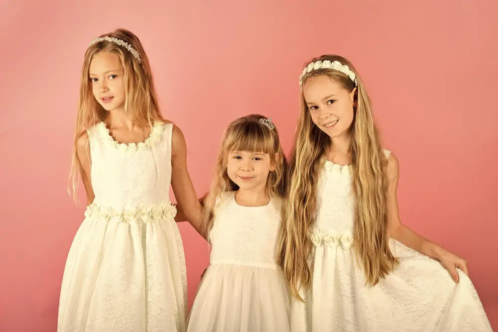 Three girls wearing white junior bridesmaid dresses in different sizes standing together