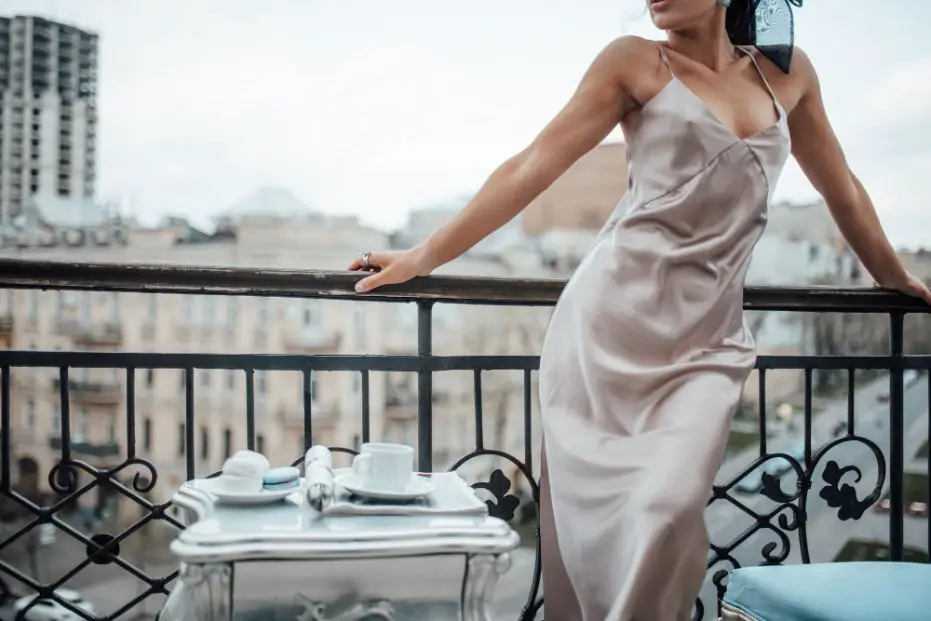 Woman wearing a silky panne satin slip dress while leaning on a balcony railing in an urban setting