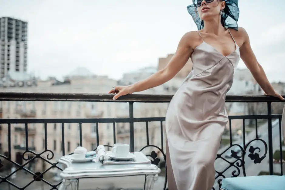 Woman wearing a silver charmeuse slip dress while standing on a balcony with a city view