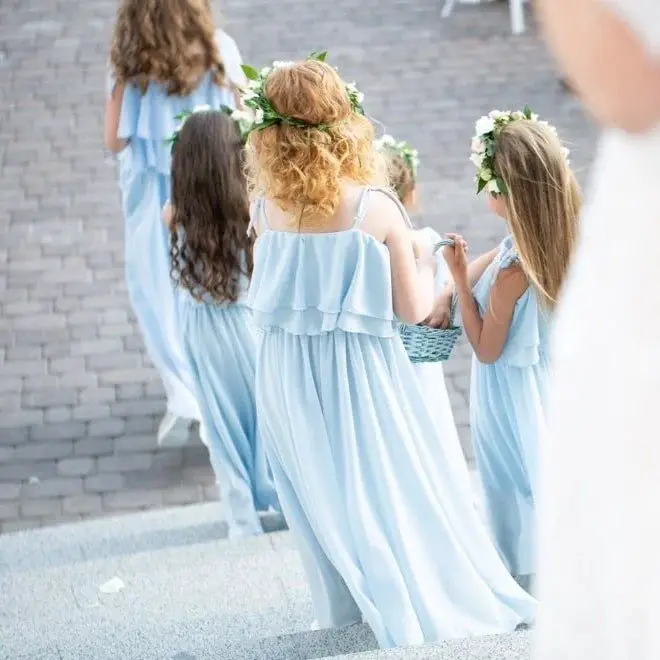 Junior bridesmaids walking together in light blue chiffon dresses with floral crowns at an outdoor wedding