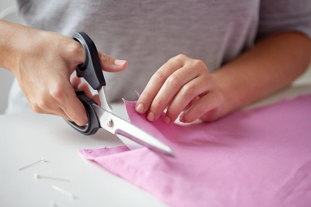 Hands cutting pink pearl chiffon fabric with scissors on a sewing table, showing careful handling of lightweight slippery material