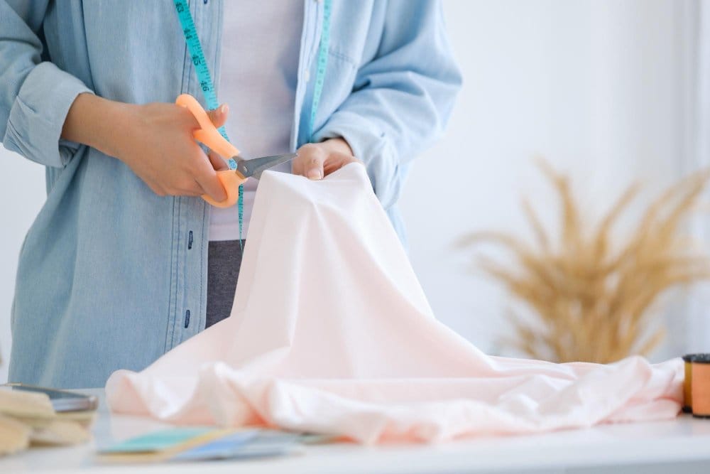 Hands cutting pale pink polyester chiffon fabric with scissors and measuring tape during sewing preparation