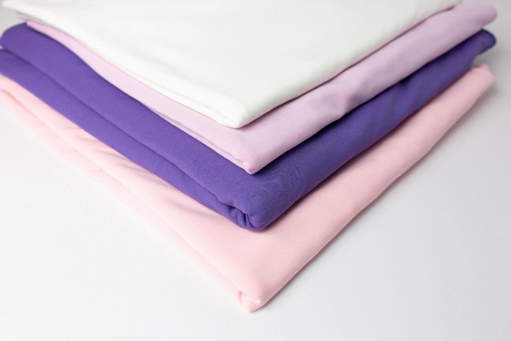 Folded stack of pastel double face chiffon fabrics in pink, lavender, purple, and white tones arranged neatly to show soft texture and smooth finish