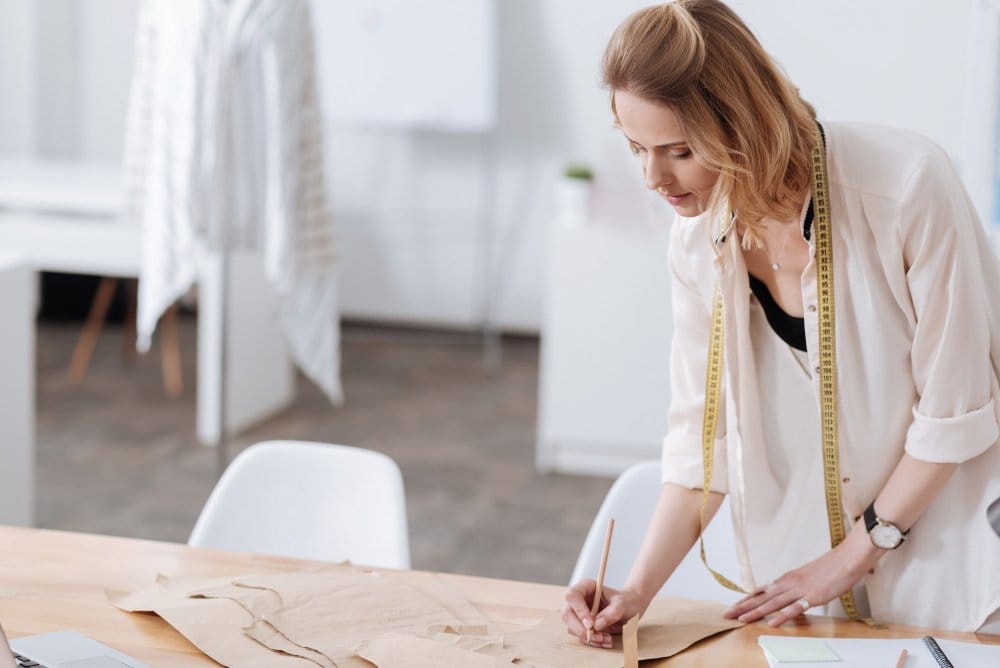 Fashion designer measuring and sketching garment patterns on a table with double face chiffon fabric nearby for dressmaking and creative styling