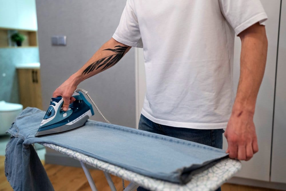 Caring for washed denim at home by ironing jeans carefully after washing. Person ironing light washed denim jeans at home on an ironing board for denim care and maintenance.