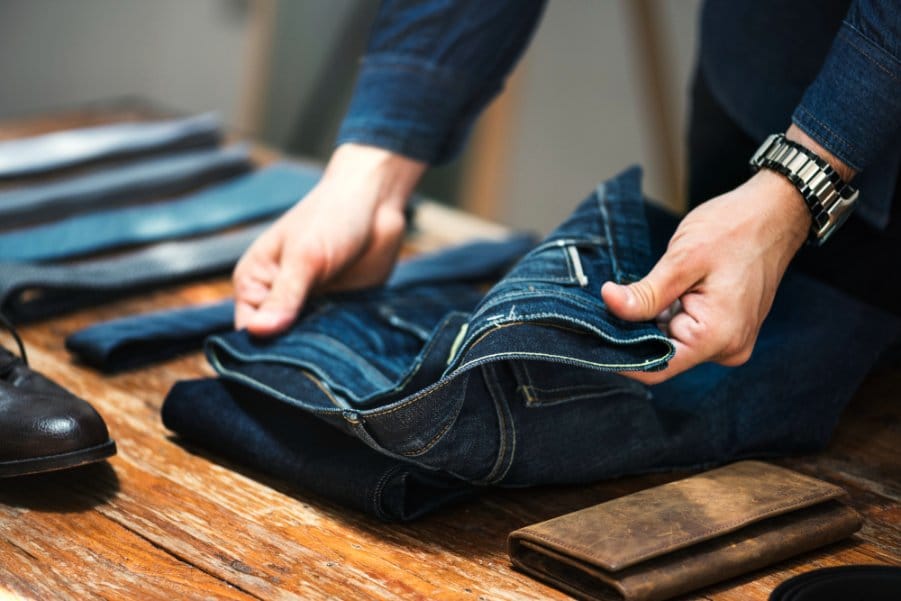 Hands folding raw denim jeans on a wooden table, representing proper raw denim care and handling.