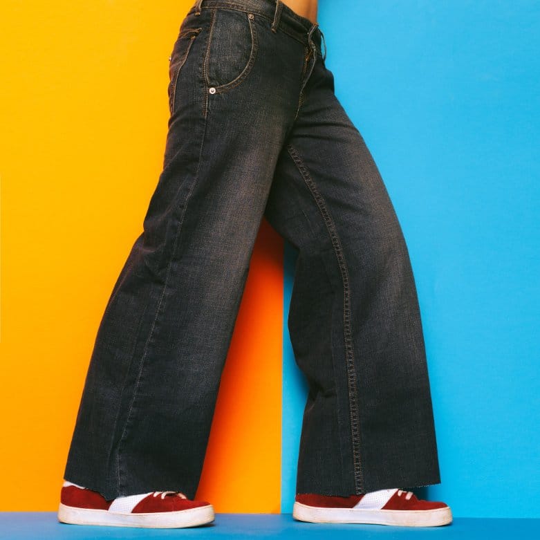 Wide-leg dark raw denim jeans styled with sneakers against a bold yellow and blue background.