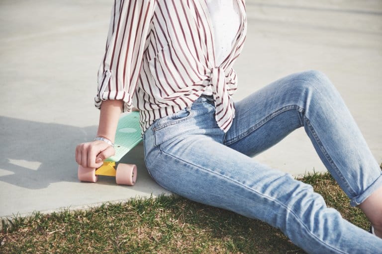 Light washed denim jeans styled in a casual outdoor look that reflects washed denim trends in 2026. Woman wearing light washed skinny jeans and a striped tied shirt while sitting outdoors with a skateboard.
