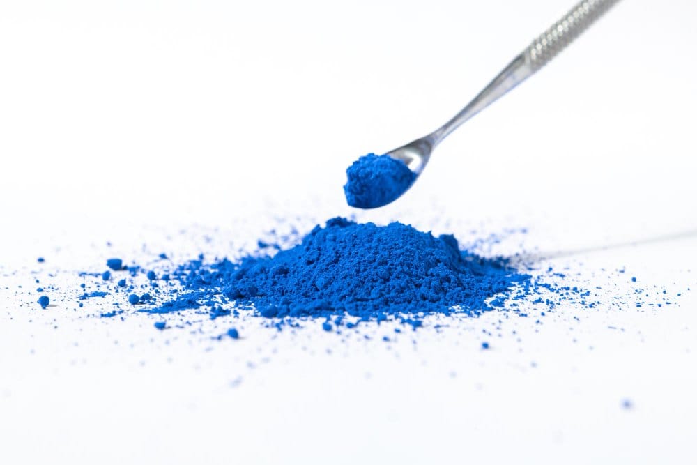 Bright blue indigo dye powder on a white surface with a metal spoon, representing the pigment used to color denim fabric.